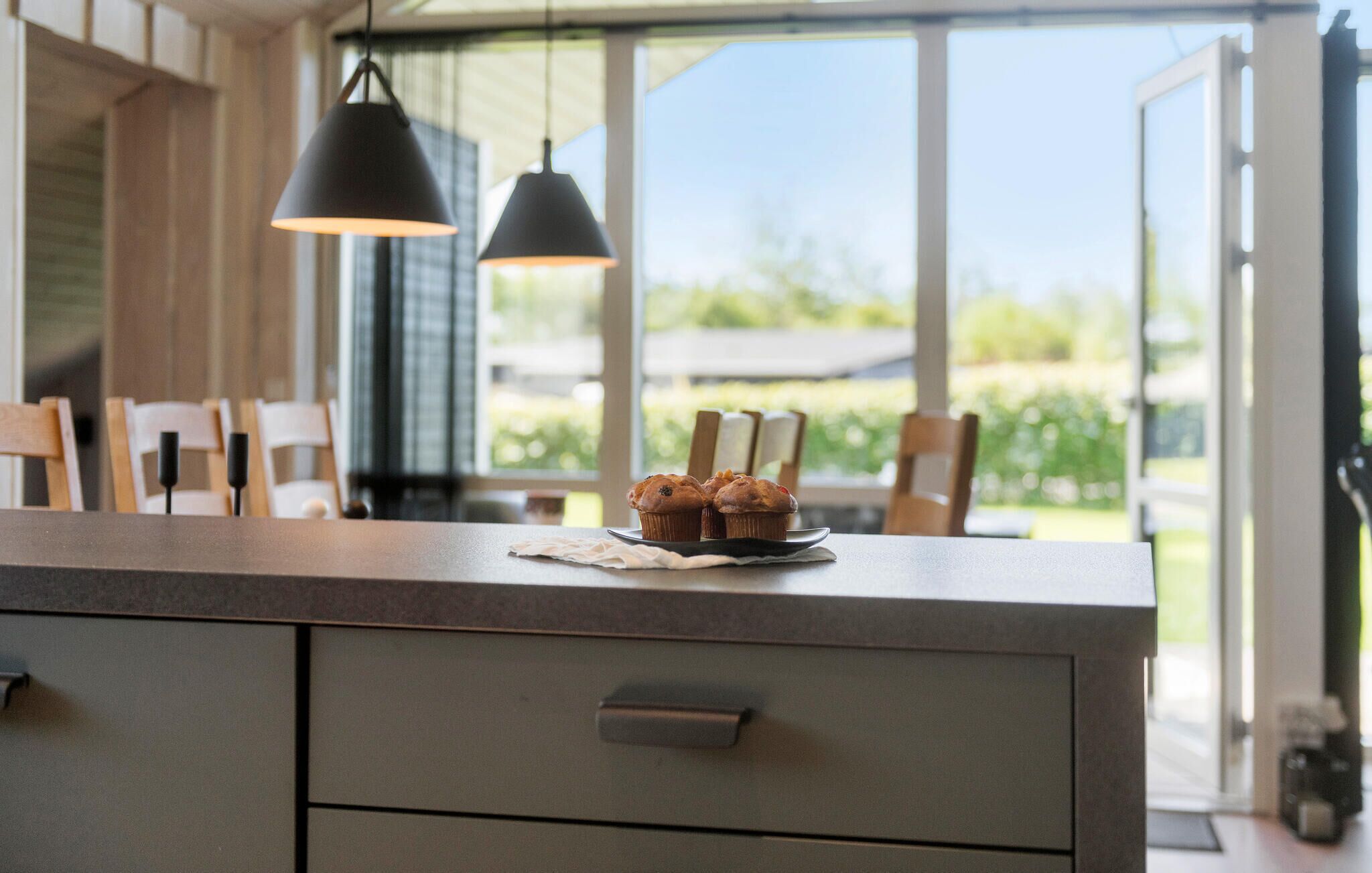 Une cuisine dotée d’une grande fenêtre, d’un plan de travail sur lequel sont disposés des muffins, et d’un luminaire suspendu au design moderne.