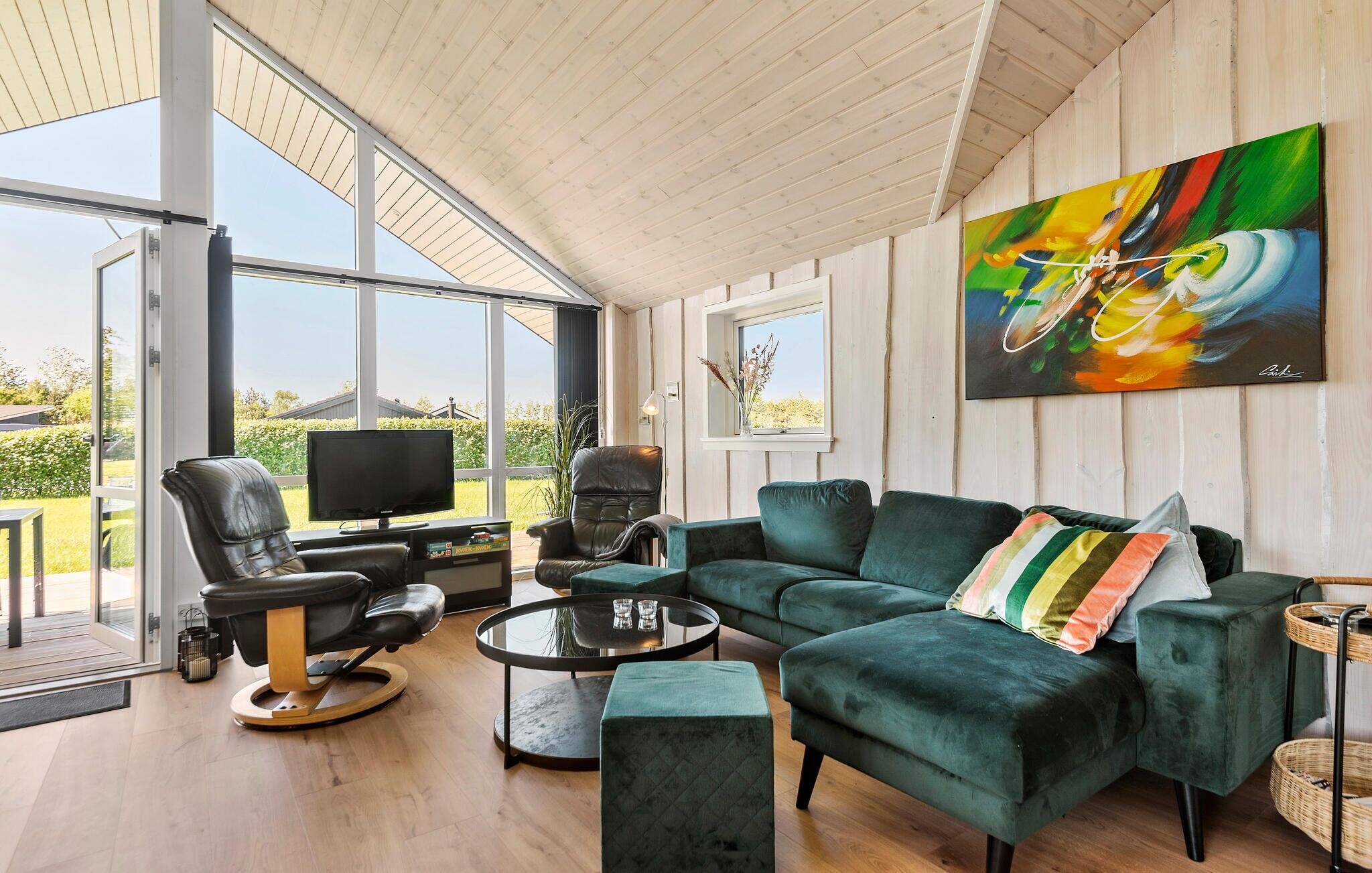 Een moderne woonkamer met een groene bank, een leren fauteuil, een televisie op een houten dressoir, en een schilderij aan de muur.