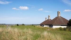 A traditional building with a tiled roof and chimneys, situated in a grassy field with a clear sky and distant water body.