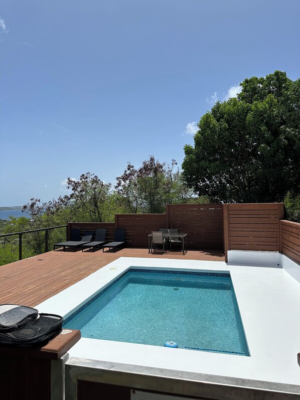 A rectangular swimming pool with a white edge, surrounded by a wooden deck. There are two lounge chairs and a table with chairs on the deck.