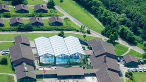 Vue aérienne d’un ensemble de bâtiments avec une piscine centrale, entouré de plusieurs bâtiments plus petits, d’un parking et de pelouses verdoyantes.
