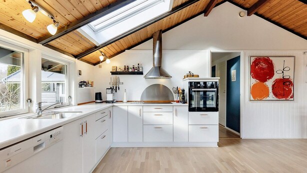 Una cocina moderna con armarios blancos, un lavavajillas Bosch, una campana extractora de acero inoxidable y un ventanal grande sobre el fregadero.