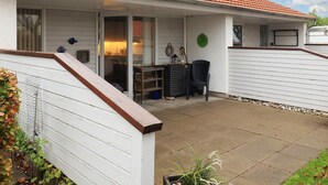 Un patio abrité avec une table et des chaises, une plaque décorative verte accrochée au mur et un bâtiment blanc surmonté d’un toit en tuiles rouges.