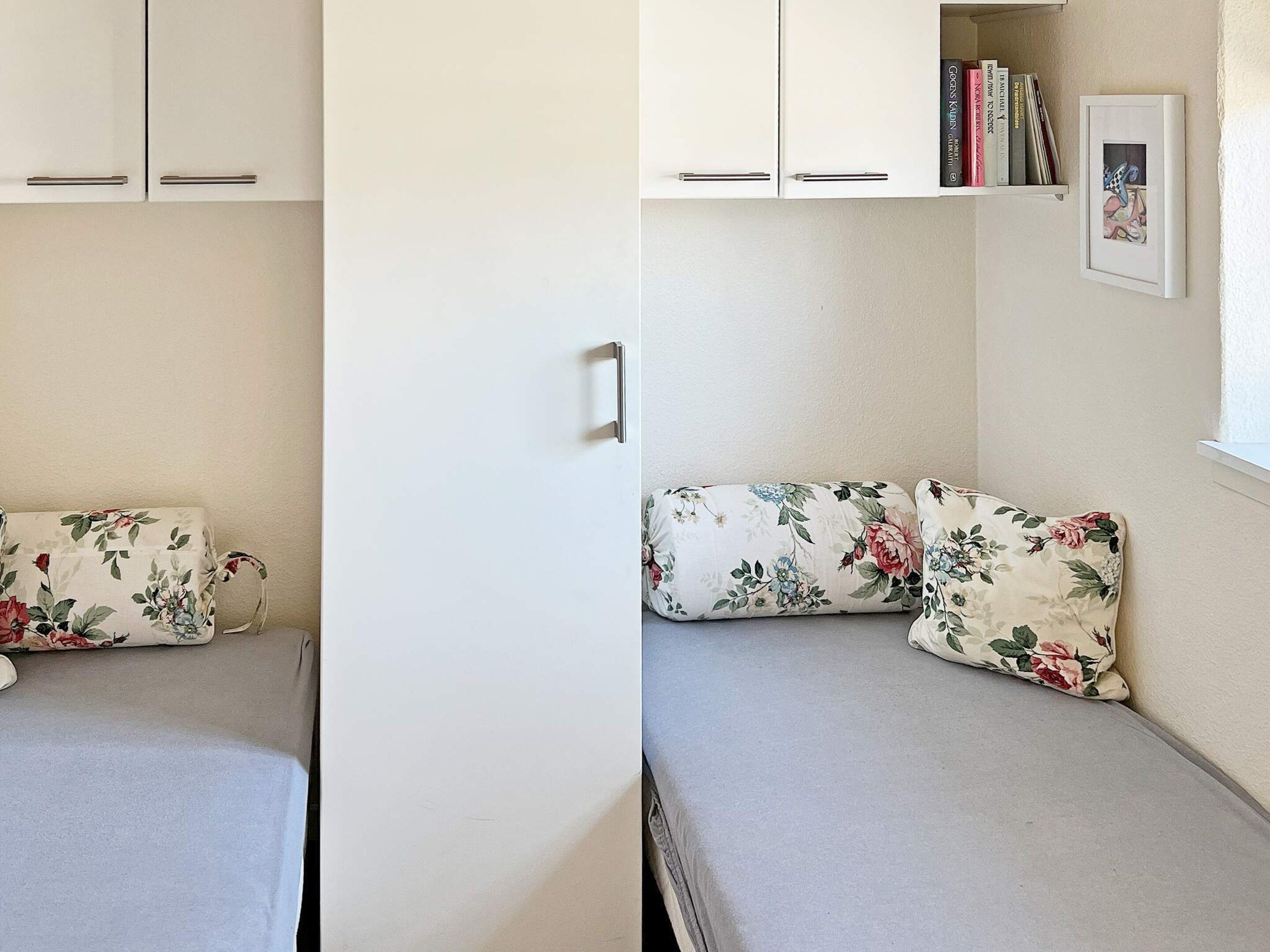 Een kleine, compacte kamer met een bed, een nachtkastje, een plank met boeken en een ingelijst schilderij, en een raam.