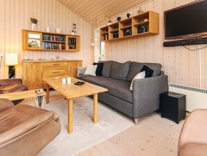 En hyggelig stue med en grå sofa, et træbord, og læderlænestole. Der er en reol med bøger og pyngeting, et fladskærms-tv og et træskab.