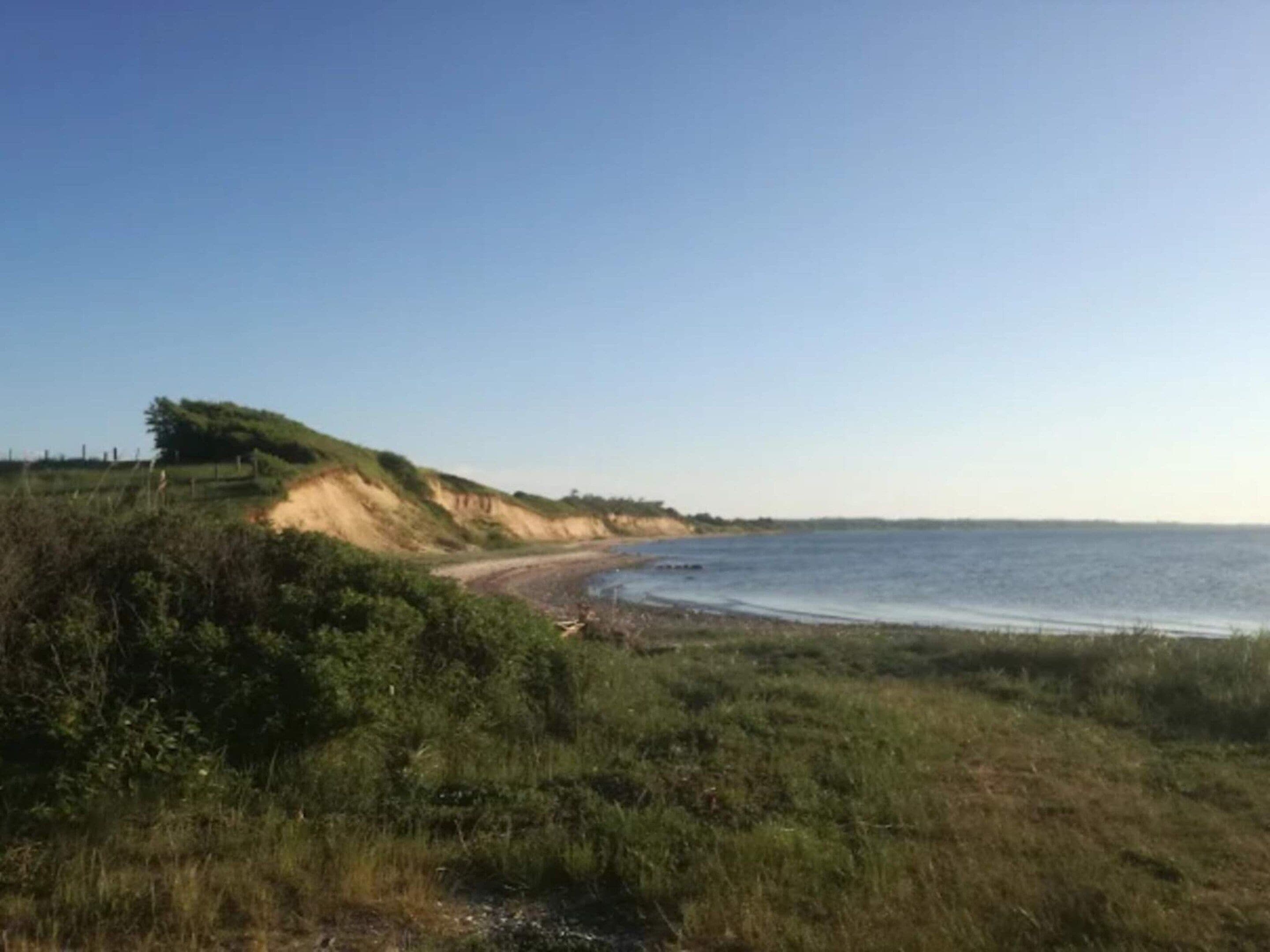 Paisagem costeira com uma praia de areia, dunas gramadas e céu claro.