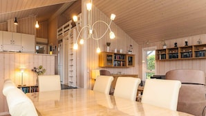 A spacious dining area with a wooden table and chairs, a kitchen in the background, and a bookshelf filled with books.