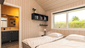 A bedroom with two beds, a wooden ceiling, a window with a view of greenery, a bookshelf, and a kitchen area visible through an open door.