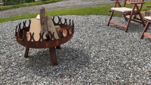A round wooden fire pit with a metal grill, surrounded by gravel, with a wooden chair and a view of a hilly landscape under a cloudy sky. - Apartment in St. Stefan im Rosental (Sankt Stefan im Rosental)