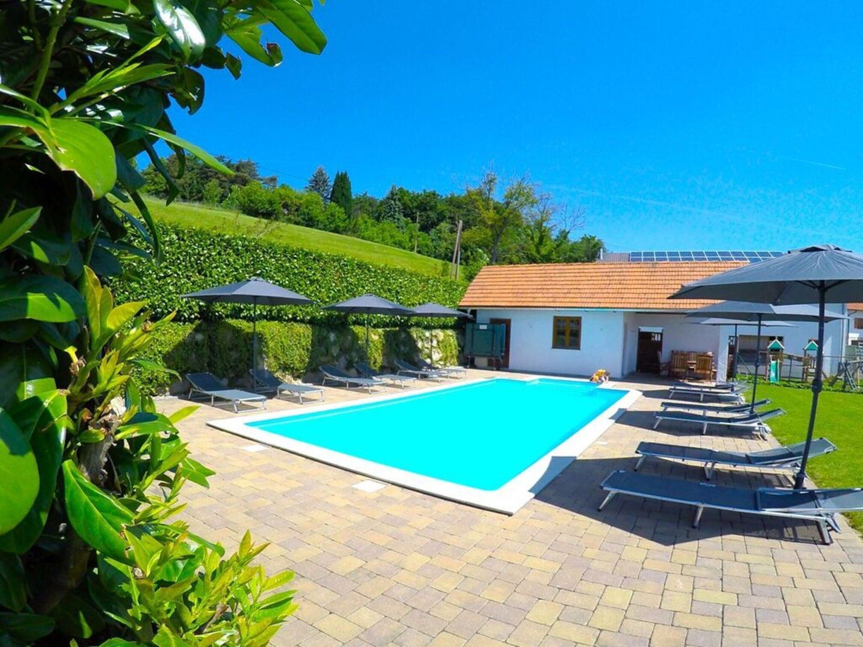A swimming pool with lounge chairs and umbrellas, surrounded by greenery and a building with a tiled roof.