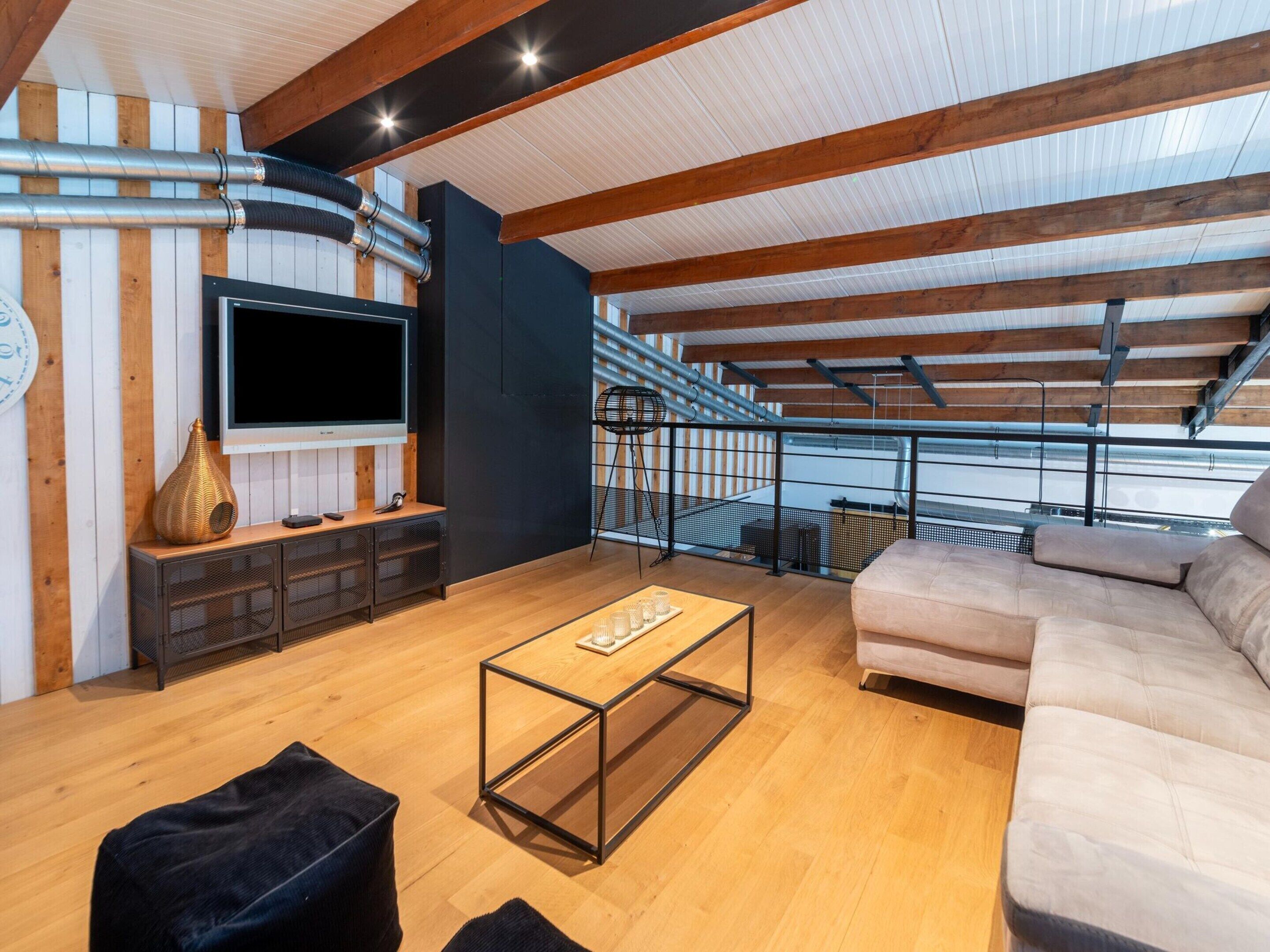 Un ampio loft con soffitto alto, travi in legno a vista, un'ampia televisione a schermo piatto, un divano, un tavolino da caffè e una scala che conduce a un piano superiore.