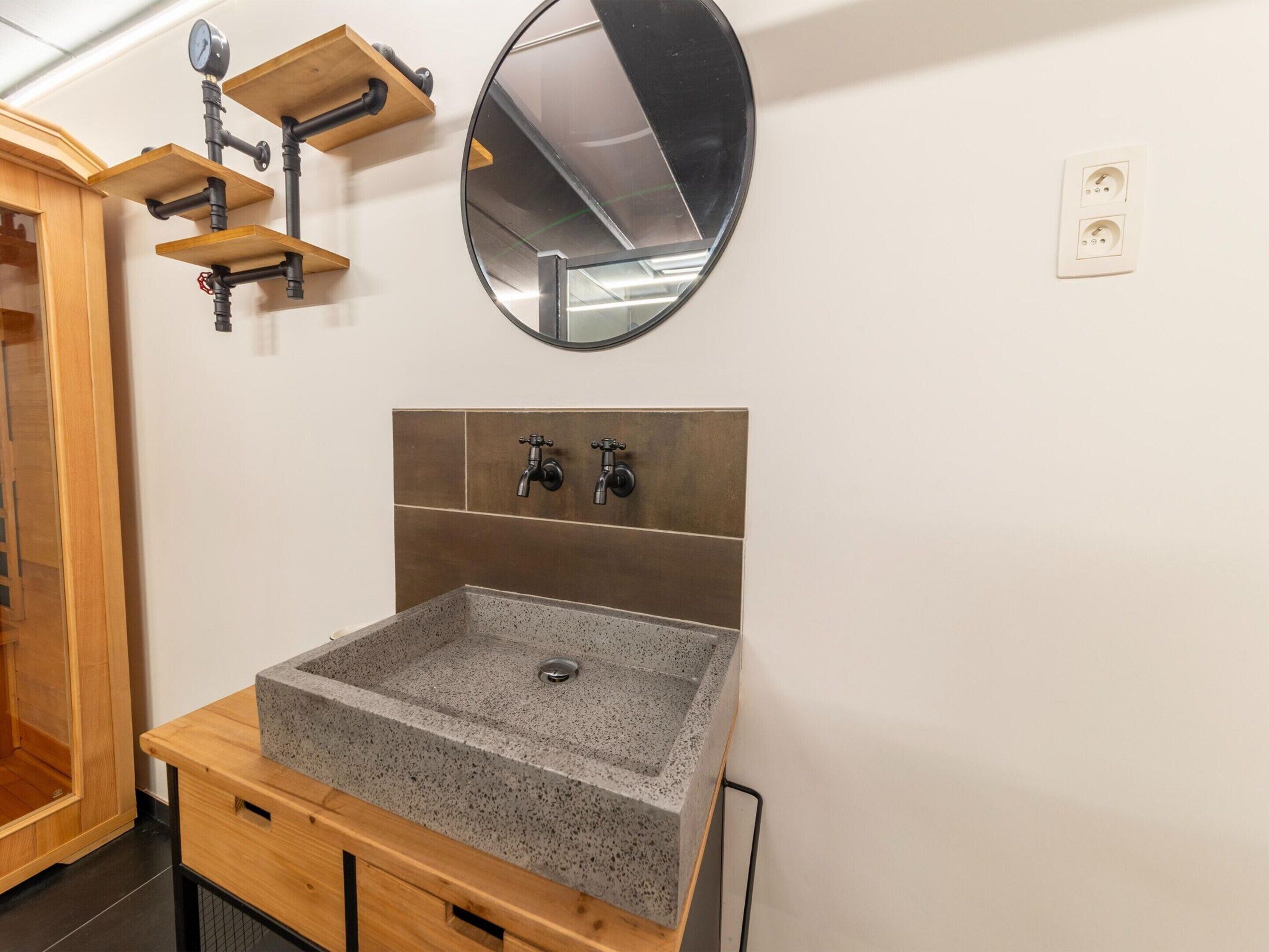 Un baño con lavamanos de granito, estantes de madera, un espejo redondo y un perchero para toallas.
