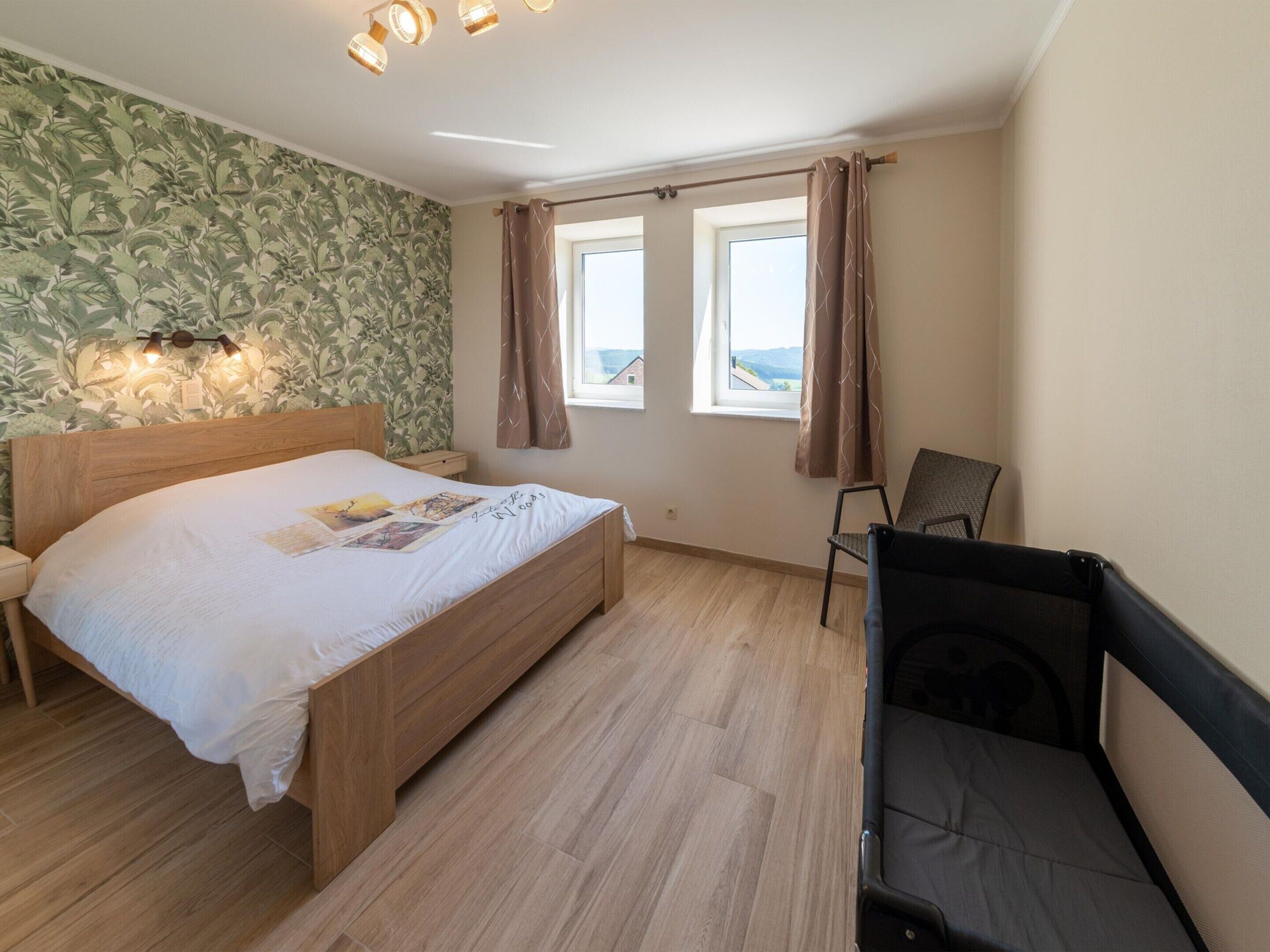 Ein Schlafzimmer mit einem hölzernen Bett, zwei mit Vorhängen versehenen Fenstern, einem Sessel und einer Wand mit Blättermotiv.