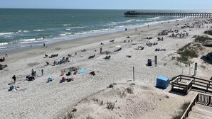 Una playa con un muelle que se adentra en el océano, terreno arenoso y algunas personas.
