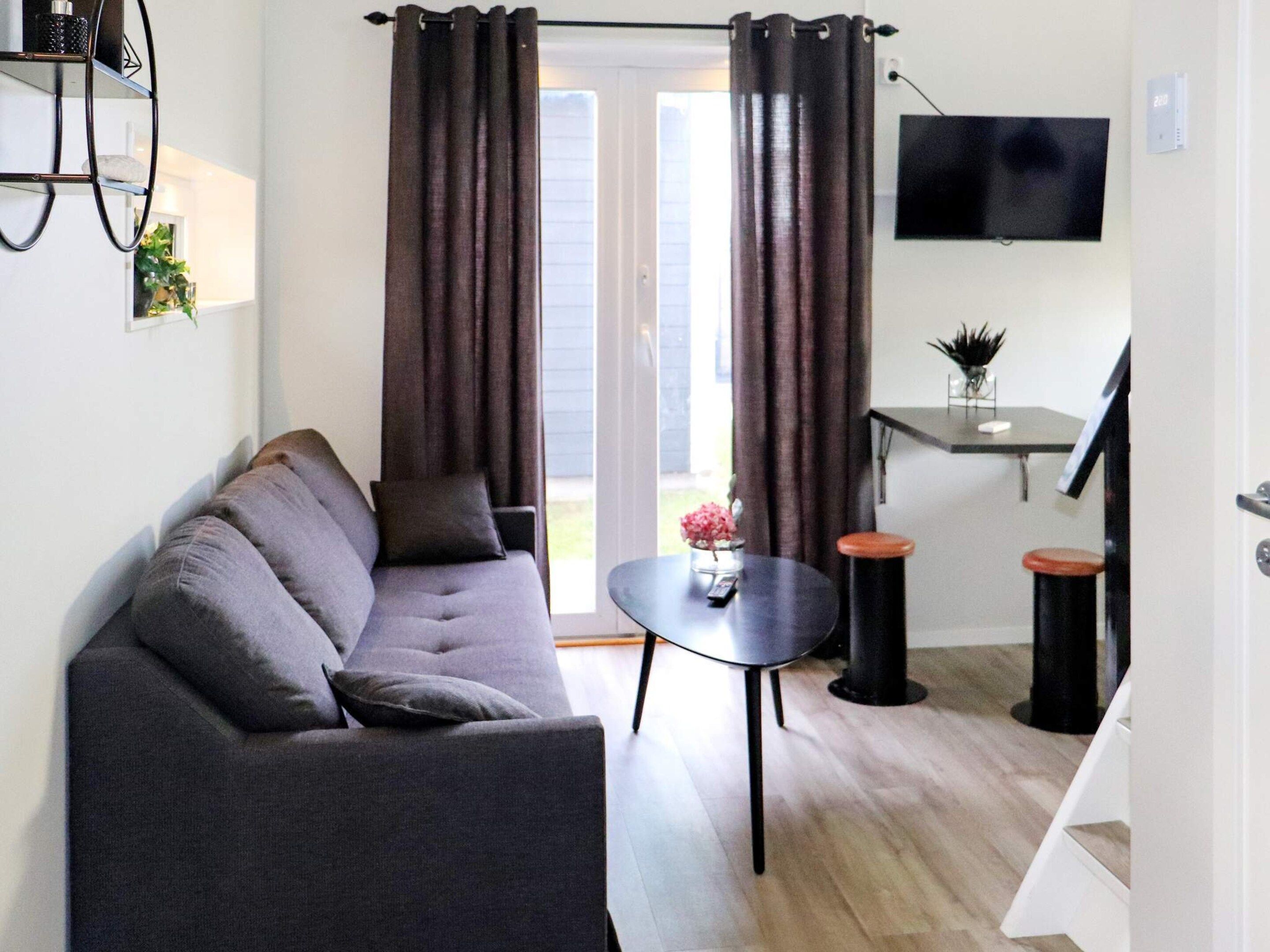 A modern living room with a grey sofa, a small round table, and two stools. There is a TV mounted on the wall and a plant on a shelf.