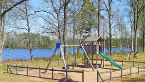 A playground with a swing set, a slide, and a small wooden structure, surrounded by a fence and a lake in the background. - 6 Person Holiday Home in Tingsryd-by Traum (Tingsryd)