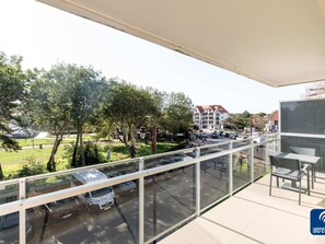 A balcony with a glass railing overlooking a parking area, a building with a red roof, and a grassy area with trees. - Parkhouse B201 Cosy Apartment in De Haan (De Haan)