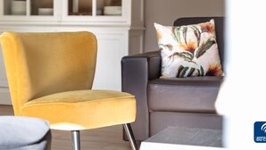 Un fauteuil jaune et un canapé foncé, agrémenté d’un coussin à motifs floraux, se trouvent dans une pièce où l’on aperçoit, à l’arrière-plan, une armoire blanche.