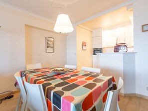 Un coin repas avec une table recouverte d’une nappe colorée, des chaises blanches, un bol de collations, une radio et, au fond, une cuisine.