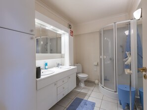 Une salle de bain avec un lavabo blanc, un miroir et des toilettes, une douche avec une porte en verre et un tapis bleu sur le sol.