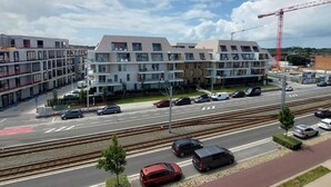 Ein modernes, mehrstöckiges Wohngebäude mit Balkonen, einem Parkplatz und einem Gleis, das parallel zur Straße verläuft.