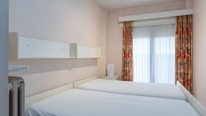 Ein Hotelzimmer mit zwei Betten, einem Fenster mit Vorhängen, einem Nachttisch und einem an der Wand befestigten Regal.
