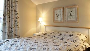 Una cama bien hecha con estampado de velero, una lámpara de noche y dos cuadros en la pared.