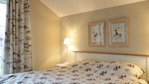 Ein Schlafzimmer mit einem Bett, Vorhängen, einem Nachttisch, einer Lampe und zwei gerahmten Bildern an der Wand.