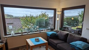 A cozy living room with a large window, a wooden table, a sofa, and a radiator.