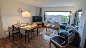 A modern living room with a dining area, a sofa, a TV, and a large window with a view of buildings.
