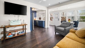 A modern living room with a flat-screen TV, a sofa, a chair, and a wooden shelf with books and a bag.