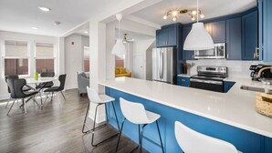 A modern kitchen with blue cabinets, white countertops, and stainless steel appliances. There is a dining area with a table and chairs, and a yellow accent wall.