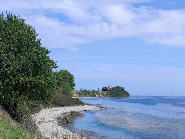 Un paisaje costero con una playa de guijarros, abundante vegetación y una colina distante con construcciones.