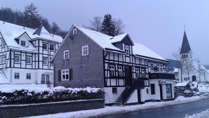A traditional half-timbered building with a steep roof, a small adjacent structure with a sign, and a church with a tall steeple in the background. - Holiday Home Near Winterberg Ski Slopes (Olsberg)