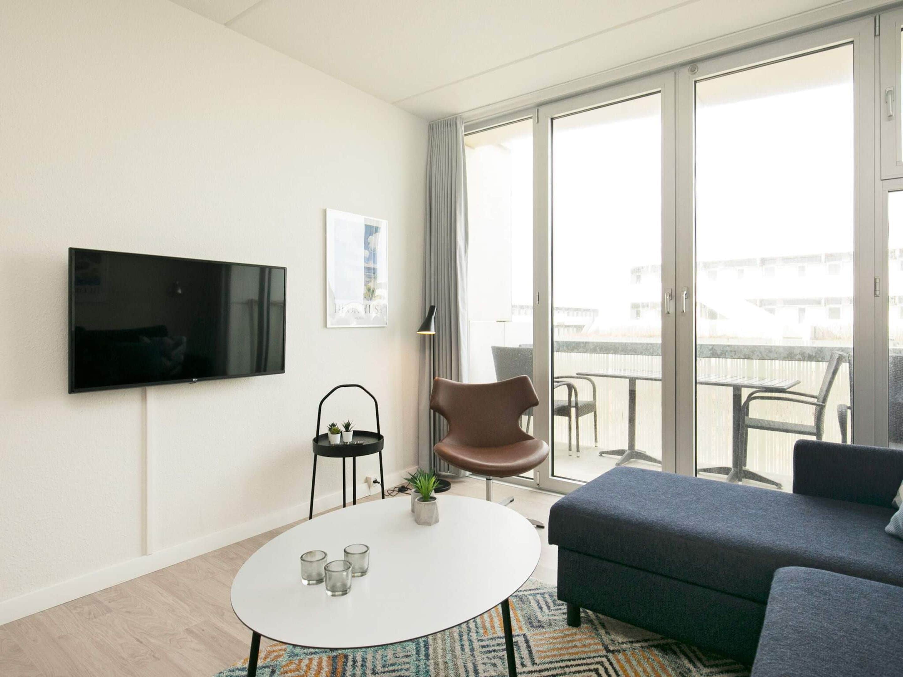 A modern living room with a flat-screen TV, a round coffee table with glasses, a brown chair, a grey sofa, and a balcony with outdoor furniture.