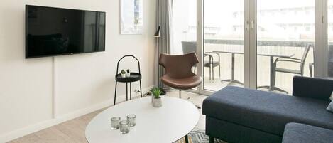 A modern living room with a flat-screen TV, a round coffee table with glasses, a brown chair, a grey sofa, and a balcony with outdoor furniture.