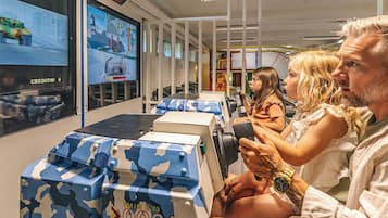 A room with a large screen displaying a video game, seating with camouflage-patterned boxes, and a person holding a game controller.