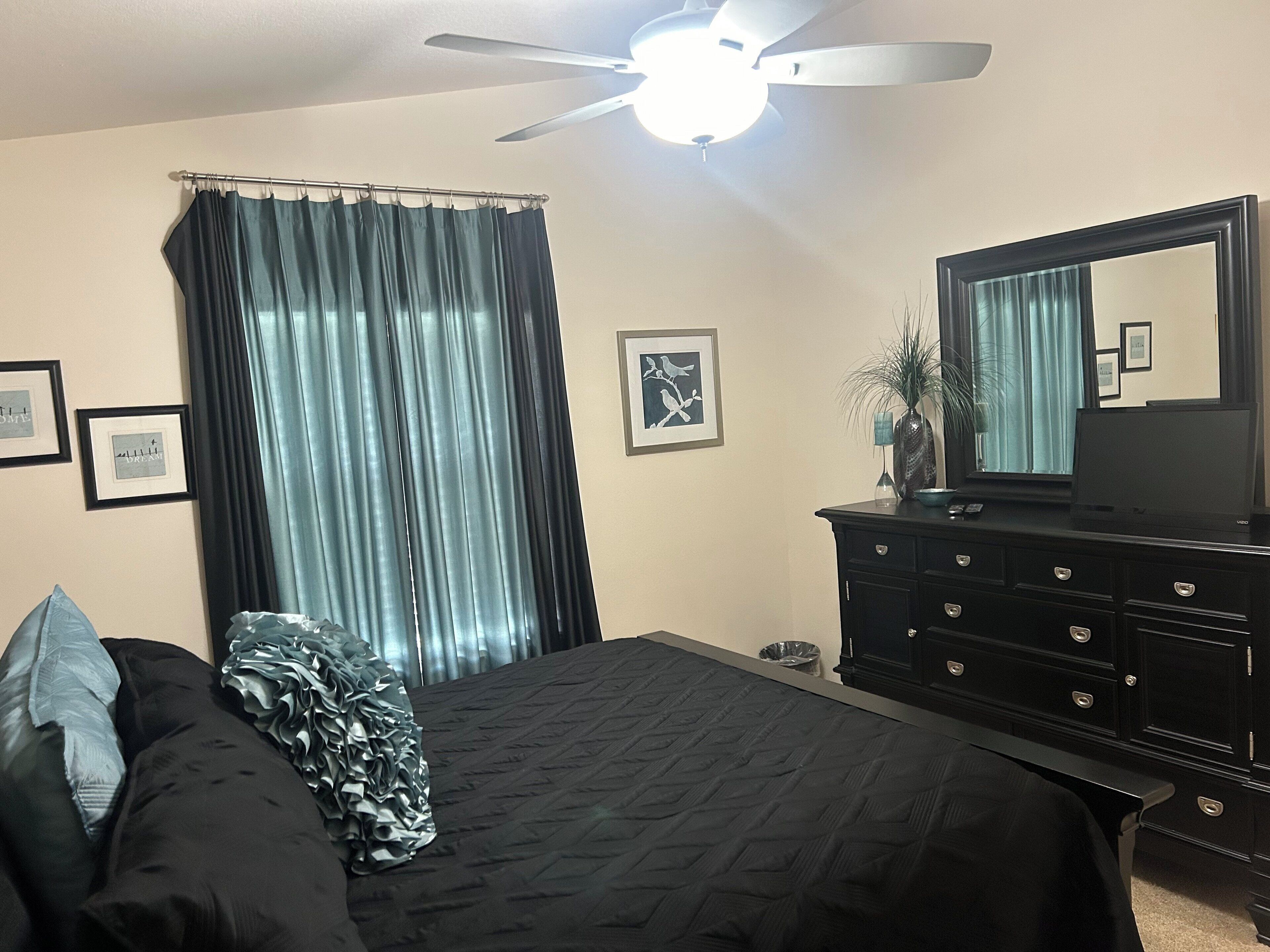 A bedroom with a bed, a dresser, a ceiling fan, and a window with curtains.