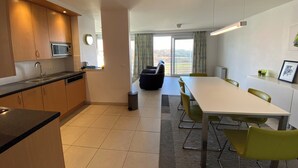 A modern kitchen with a microwave, sink, and wooden cabinets. There is a dining area with a table and chairs, and a view of the outdoors through large windows. - Apartment Nieuwpoort Near Beach & Marina (Nieuwpoort)