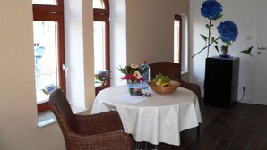 A dining area with a round table, wicker chairs, a white tablecloth, a fruit bowl, a vase with flowers, and a potted plant. - Holiday Apartment Amsterdam (Plau am See)