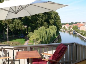 A balcony with a table and chairs, overlooking a river with trees and buildings in the background. - Holiday Apartment Amsterdam (Plau am See)