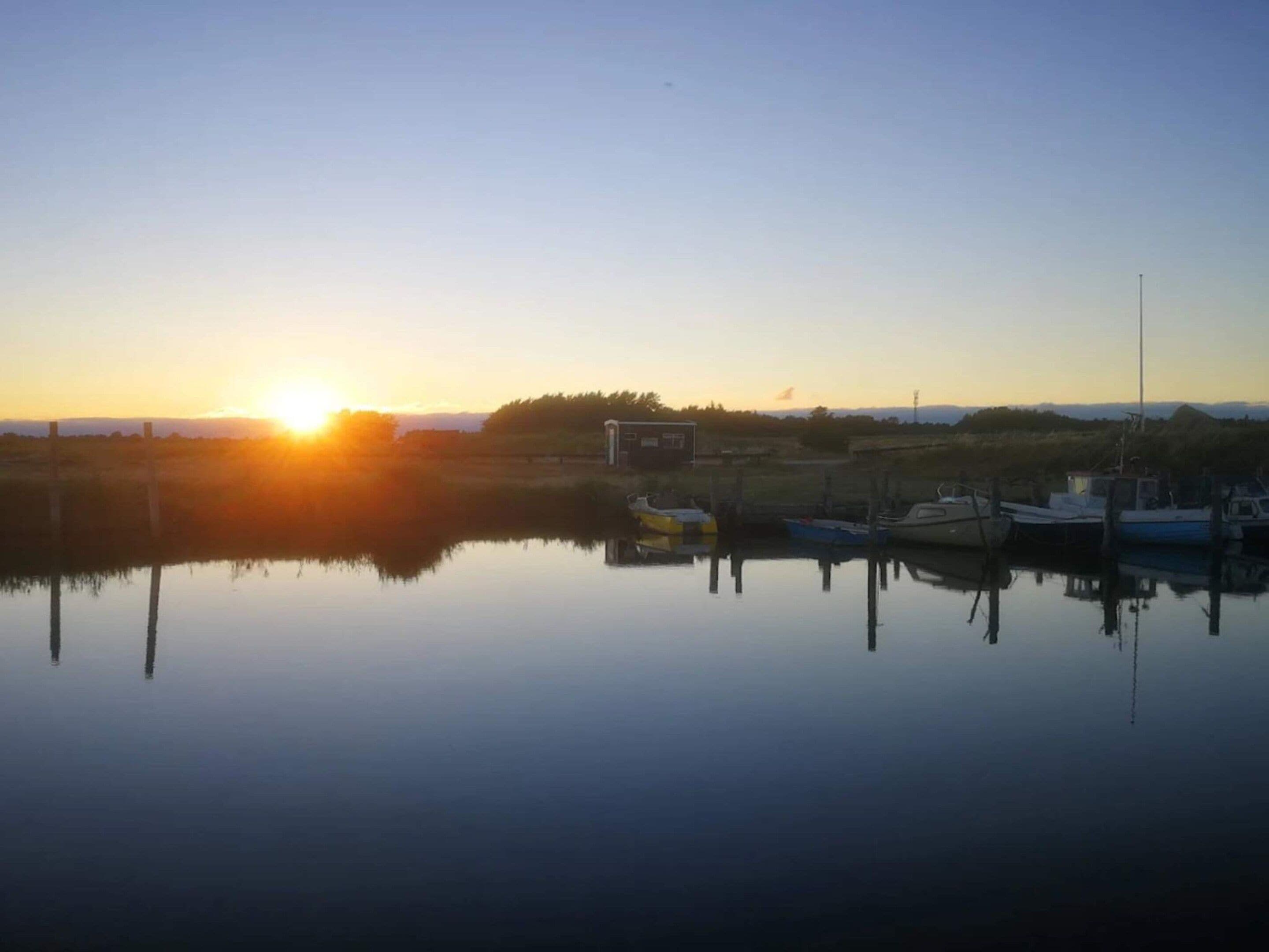 A calm body of water reflecting the sun, boats docked, and a small building in the distance.