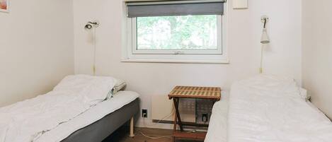 A small, single-room accommodation with two beds, a window with a view of greenery, a wooden ceiling, and a small table.