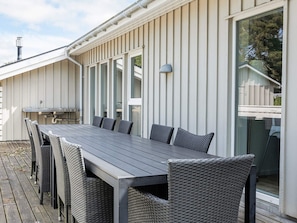 A covered outdoor dining area with a long table and wicker chairs, a grill, and a large window. - 12 Person Holiday Home in Rodby (Rodby)