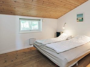Een slaapkamer met een houten plafond, een raam met uitzicht op bomen, een bed met witte lakens en een wandlamp.