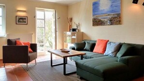 Un salon avec un canapé vert, une table basse en bois et un fauteuil noir. Il y a un tableau au mur, un radiateur et une porte qui donne sur l’extérieur.