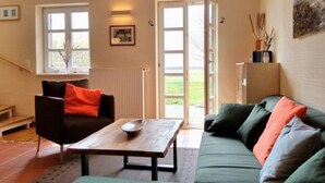 Un salon avec un canapé, une table et une chaise. Il y a une fenêtre et une porte qui donne sur l’extérieur.