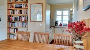Un coin repas avec une table et des chaises en bois, une bibliothèque remplie de livres, une carte accrochée au mur et un espace cuisine avec une fenêtre.