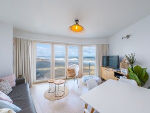 A modern living room with a large window, a sofa, a dining table, and a TV. - Cozy Studio With Sleeping Area and sea View (Middelkerke)