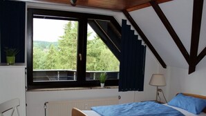 Une chambre avec une grande fenêtre, un lit avec une literie bleue, une table de chevet, une lampe et une vue sur la verdure à l’extérieur.